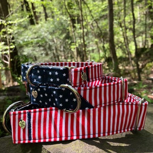 May include: A stack of three dog collars with patriotic designs. One collar features a navy blue background with white stars, while the others have red and white stripes. Each collar has a silver metal D-ring and a small heart-shaped charm that says "Made with Love."
