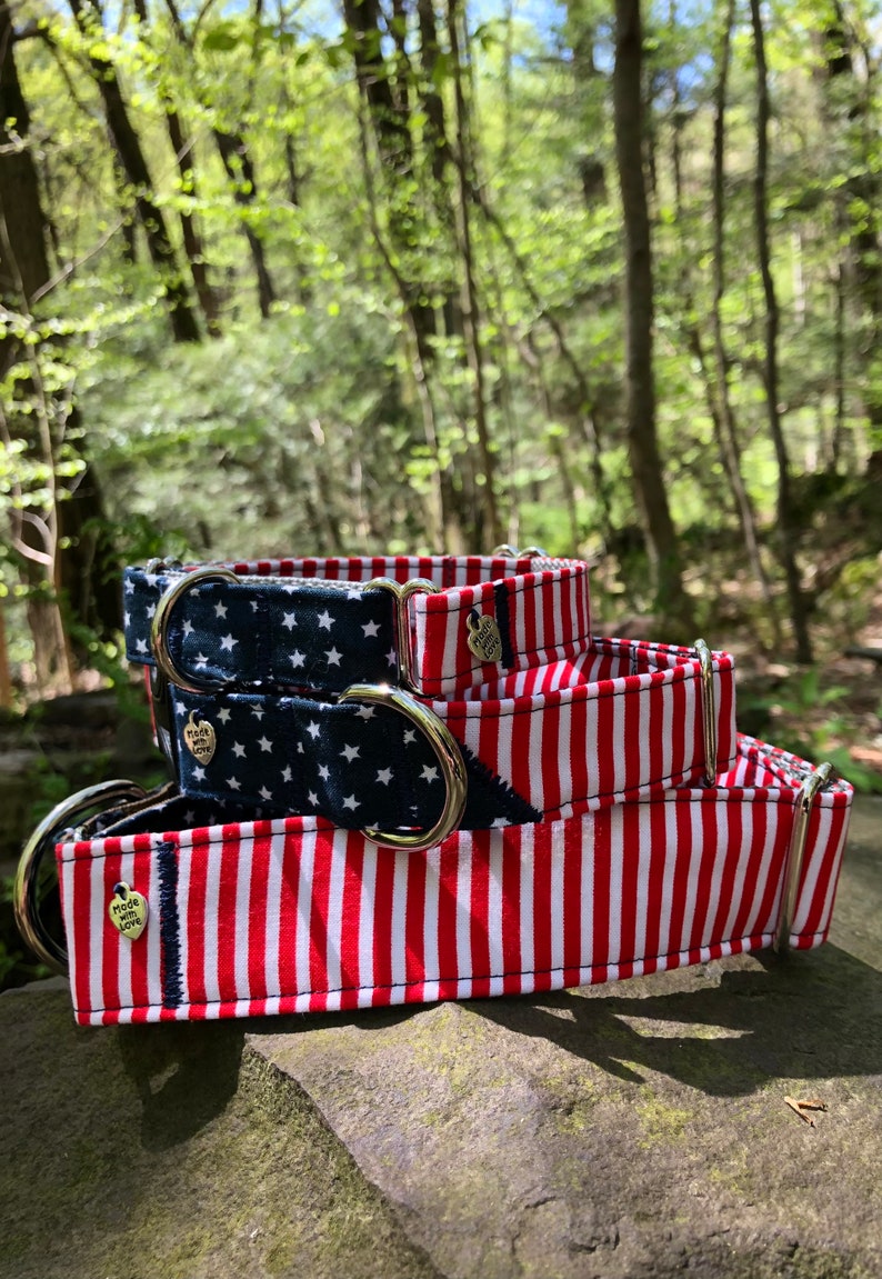 May include: A stack of three dog collars with patriotic designs. One collar features a navy blue background with white stars, while the others have red and white stripes. Each collar has a silver metal D-ring and a small heart-shaped charm that says "Made with Love."
