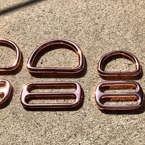 Op de afbeelding: Assortiment van roségouden metalen hardware, waaronder D-ringen, O-ringen en schuifgespen. Deze metalen onderdelen worden vaak gebruikt in handwerk en sieraden maken.