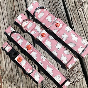 May include: Four pink dog collars with a white ghost and orange pumpkin pattern. The collars are different sizes and have different hardware, including a buckle, a D-ring, and a slider.