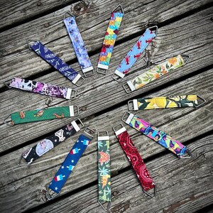 May include: A collection of 12 colorful fabric wrist straps with different patterns, including lemons, flowers, dinosaurs, and a lobster. Each strap has a silver metal loop at each end.