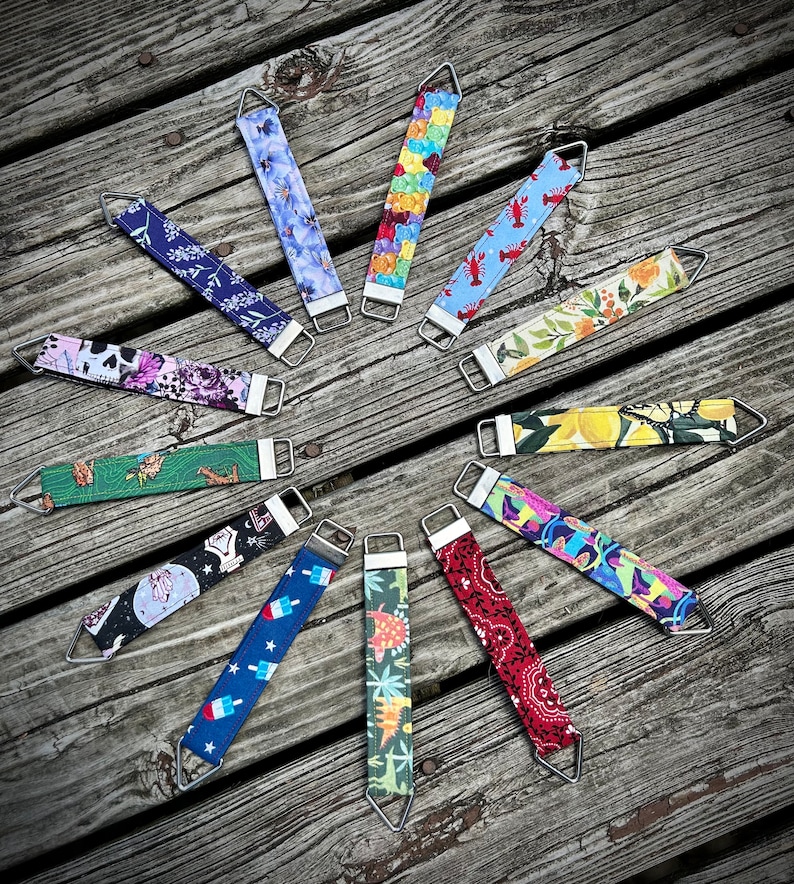 May include: A collection of 12 colorful fabric wrist straps with different patterns, including lemons, flowers, dinosaurs, and a lobster. Each strap has a silver metal loop at each end.