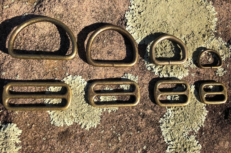 May include: Assortment of eight brass metal hardware pieces, including D-rings and rectangular buckles, arranged on a rocky surface with green lichen.