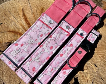 May include: Three pink dog collars with a pig print design. The collars are made of fabric and have silver metal hardware. The collars are arranged in a row, with the largest collar at the top and the smallest collar at the bottom.