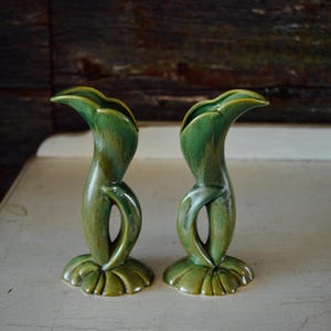 May include: Two green ceramic bud vases with a unique, organic design. The vases have a curved, elongated shape with a flared top and a base resembling leaves. The green glaze has variations in tone, adding depth and visual interest. These vases are ideal for displaying single stems or small floral arrangements.