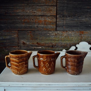 May include: Three brown ceramic creamers with handles and spouts. The left and right creamers feature a horizontal chevron pattern, while the center creamer displays a geometric diamond and border design. Each piece has a glossy finish.