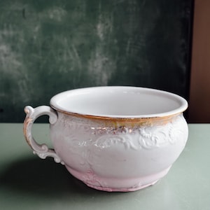 Op de afbeelding: Een witte en roze keramische kom met een handvat en sierlijke details. De rand is voorzien van een gouden band. De kom staat tegen een gedempte groene achtergrond.