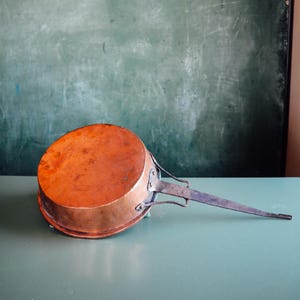 May include: A copper saucepan with a long, flat handle. The pan has a rounded base and a bright, reflective surface. The handle is made of a darker metal and is attached to the pan with rivets. The pan is resting on a light green surface.