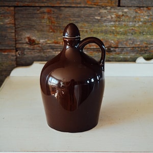 May include: A brown ceramic jug with a lid and a handle. The jug has a glossy finish and is sitting on a white surface.