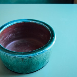 May include: A small, round ceramic bowl with a teal exterior and a maroon interior. The bowl has a glossy finish and sits on a teal surface. The rim of the bowl is a lighter shade of teal.