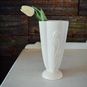 May include: A cream-colored ceramic vase with a tulip design, holding a single yellow tulip. The vase has a flared shape and a pedestal base. The background is a rustic wooden wall.