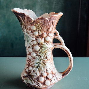 Può includere: Brocca in ceramica con manico, decorata con un motivo a uva e foglie. La brocca presenta una combinazione di colori marrone e crema, con un'ampia apertura e un manico curvo. La brocca è alta circa 20 cm.