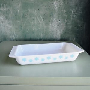 May include: A rectangular white baking dish with a light blue floral pattern along the bottom edge. The dish is empty and sits on a light green surface. The background is a dark green chalkboard.
