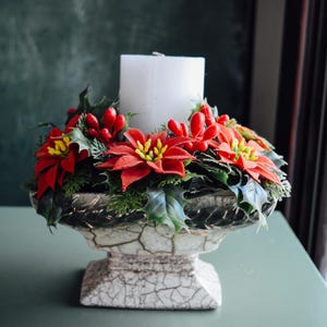 Pode incluir: Uma vela branca está no centro de um vaso de cerâmica, cercada por um anel de flores de poinsettia vermelhas e folhas de azevinho verdes.