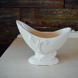 Puede incluir: Una salsera de cerámica blanca con base de pedestal y forma curva, similar a un barco. El exterior presenta diseños florales en relieve. La salsera se exhibe sobre una superficie blanca contra un fondo de madera rústica.