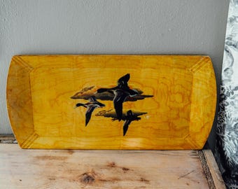 May include: A yellow wooden tray featuring a black and white image of ducks in flight against a cloudy sky. The tray has a slightly rounded edge and a wood grain pattern.
