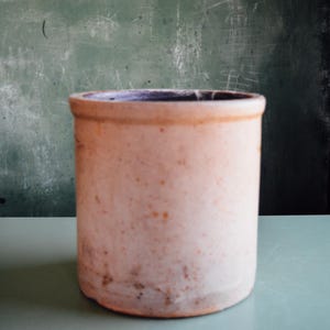 Pode incluir: Um pote de grés cilíndrico com um exterior bege claro salpicado e uma borda lisa e arredondada. O interior do recipiente é escuro e a superfície externa apresenta variações subtis de cor e textura ao longo das suas paredes verticais.