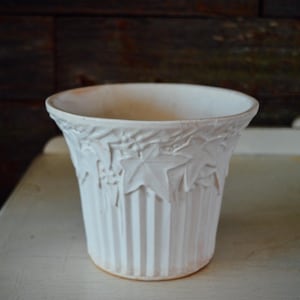 Pode incluir: Um vaso de cerâmica branco com um design canelado e um padrão de folhas em relevo. O vaso tem uma abertura larga e uma forma ligeiramente alargada. O fundo é uma parede de madeira rústica e uma superfície branca.