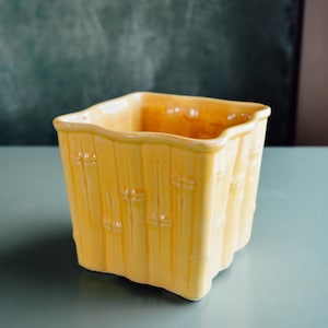 May include: A square, yellow ceramic container with a bamboo-like texture. The container has a wavy top edge and is sitting on a light blue surface. The background is a dark green chalkboard.