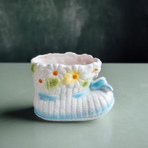 Könnte beinhalten: Ein weißer Baby-Stiefel-Blumentopf aus Keramik mit blauer Schleife und Besatz. Der Stiefel ist mit Gänseblümchen und orangefarbenen Akzenten verziert. Der Topf hat ein strukturiertes, gestricktes Aussehen.