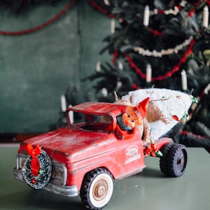 Puede incluir: Un camión de juguete rojo vintage con un árbol de Navidad en la parte trasera y una corona en la parte delantera. Una figura de elfo está en el asiento del conductor. El camión tiene la palabra "Tonka" en el lateral.