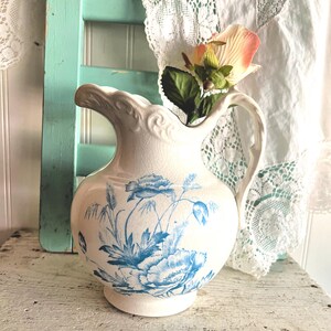 May include: A white ceramic pitcher with blue floral designs and a handle. The pitcher has a decorative rim and is placed on a distressed wooden surface. A faux flower with pink and white petals is inside the pitcher.