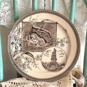 May include: Cream-colored decorative plate with sepia-toned illustrations. The plate features a central image of a lion and text that reads "LION OF LUCERNE". Floral and geometric border designs surround the central image.