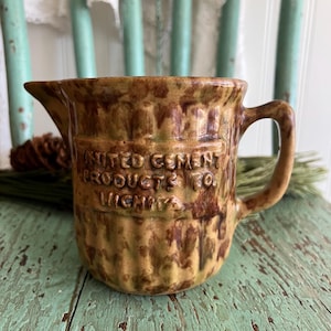 May include: Vintage brown and tan ceramic pitcher with a handle and spout. The pitcher has the text "UNITED CEMENT PRODUCTS CO. WIGHTS" embossed on the front. The pitcher is sitting on a distressed, light blue surface.