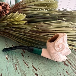 May include: A cream-colored tobacco pipe with a ram's head design. The pipe has a black stem with a green band and rests on a distressed, turquoise surface. Dried greenery and a pine cone are in the background.