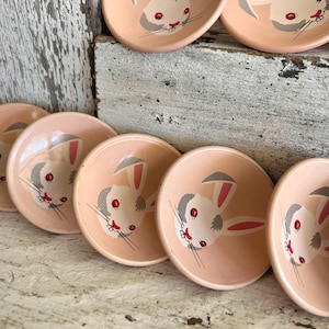 May include: A collection of small, pink ceramic bowls. Each bowl features a hand-painted bunny face with gray ears, red eyes, and a red nose. The bowls are arranged on a distressed, white wooden surface.