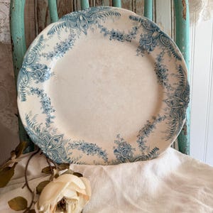 May include: A round ceramic plate featuring a cream-colored base with a decorative blue transferware pattern along the scalloped rim. The intricate design includes floral garlands, leafy sprigs, and ornate scrollwork motifs.