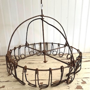 May include: A vintage, rusty metal hanging basket with a circular design. The basket features a central vertical rod with a hook at the top and a series of looped wire holders around the perimeter. The basket is designed to hang from a ceiling.