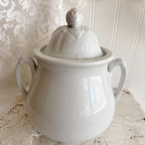 May include: A white ceramic sugar bowl with a lid and two handles. The lid has a decorative knob and a leaf-like design. The bowl is round with a glossy finish, and the handles are curved. The sugar bowl is a vintage kitchenware item.