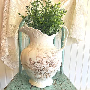 May include: A white ceramic pitcher with a floral design in brown tones, filled with green foliage and dark berries, sits on a distressed turquoise chair. The pitcher has a decorative handle and a scalloped rim. A lace curtain hangs in the background.