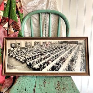 May include: A framed black and white photograph of a large gathering of people seated at long tables. The photograph is in a brown wooden frame and rests on a distressed, turquoise chair. A colorful quilt and lace curtain are in the background.