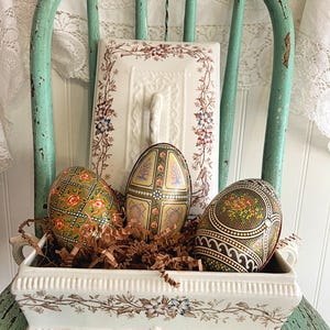 May include: An antique-style display featuring three decorative Easter eggs in a white floral-patterned tray filled with brown paper. The eggs have intricate designs in gold, green, and red. A white floral-patterned serving dish and a distressed green chair complete the scene.