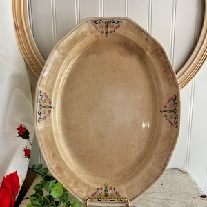 May include: An oval, antique-style serving platter with a beige surface and decorative floral accents in pink, yellow, and blue. The platter has a slightly octagonal shape and is displayed on a gold-colored stand.