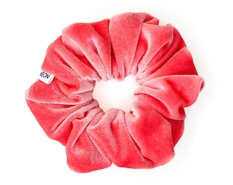 May include: A coral-colored velvet scrunchie with a soft, plush texture. The scrunchie is round with a hole in the center and a small white tag that reads "NEON". The fabric is gathered and folded, creating a voluminous appearance.