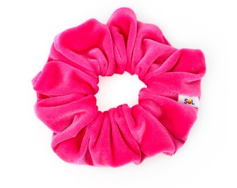 Puede incluir: Un scrunchie de terciopelo rosa brillante con una etiqueta blanca que dice "SOL" en texto negro.