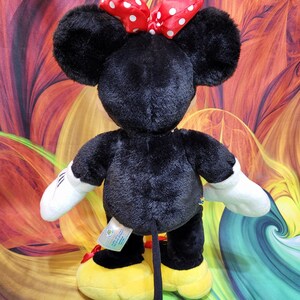 K&ouml;nnte beinhalten: Schwarze Minnie Mouse Pl&uuml;schpuppe mit gro&szlig;en schwarzen Ohren, wei&szlig;en Handschuhen und gelben Schuhen. Die Puppe hat eine rote Schleife mit wei&szlig;en Punkten auf dem Kopf. Die Puppe steht vor einem bunten Hintergrund.