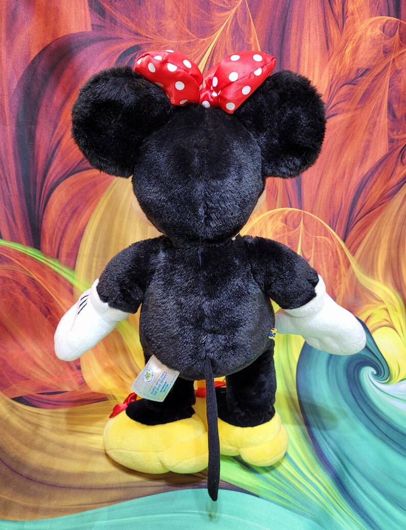 K&ouml;nnte beinhalten: Schwarze Minnie Mouse Pl&uuml;schpuppe mit gro&szlig;en schwarzen Ohren, wei&szlig;en Handschuhen und gelben Schuhen. Die Puppe hat eine rote Schleife mit wei&szlig;en Punkten auf dem Kopf. Die Puppe steht vor einem bunten Hintergrund.