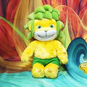 May include: A plush toy lion with a yellow body, green shorts, and a green mane. The lion has a smiling face with green eyes. The background features a colorful, abstract design with shades of red, orange, and blue.