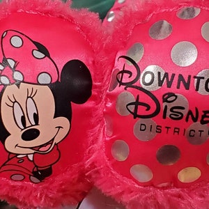 May include: Pink Minnie Mouse earmuffs with a plush trim. One side features Minnie Mouse with a polka dot bow. The other side has silver polka dots and the text "Downtown Disney District".