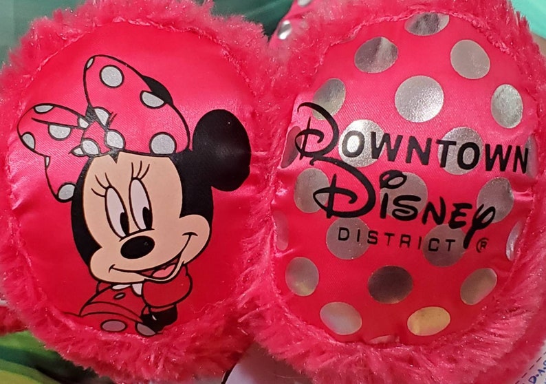 May include: Pink Minnie Mouse earmuffs with a plush trim. One side features Minnie Mouse with a polka dot bow. The other side has silver polka dots and the text "Downtown Disney District".