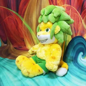May include: A plush toy lion with a yellow body, green shorts, and a green leafy mane. The lion has a smiling face with white teeth and is sitting on a teal and blue surface.