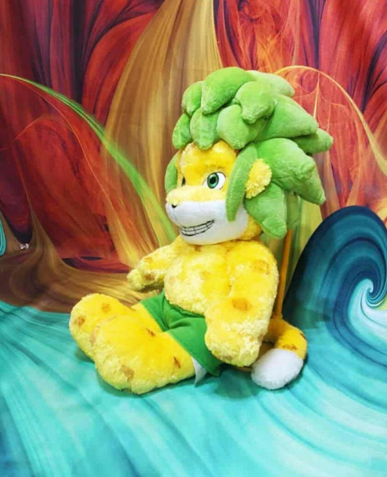 May include: A plush toy lion with a yellow body, green shorts, and a green leafy mane. The lion has a smiling face with white teeth and is sitting on a teal and blue surface.