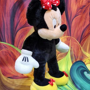 K&ouml;nnte beinhalten: Eine Pl&uuml;schpuppe von Minnie Mouse, mit einem schwarzen K&ouml;rper, wei&szlig;en Handschuhen und gelben Schuhen mit roten Schleifen. Die Puppe hat ein cremefarbenes Gesicht, schwarze Ohren und eine rote Schleife mit Punkten. Die Puppe steht vor einem bunten Hintergrund.