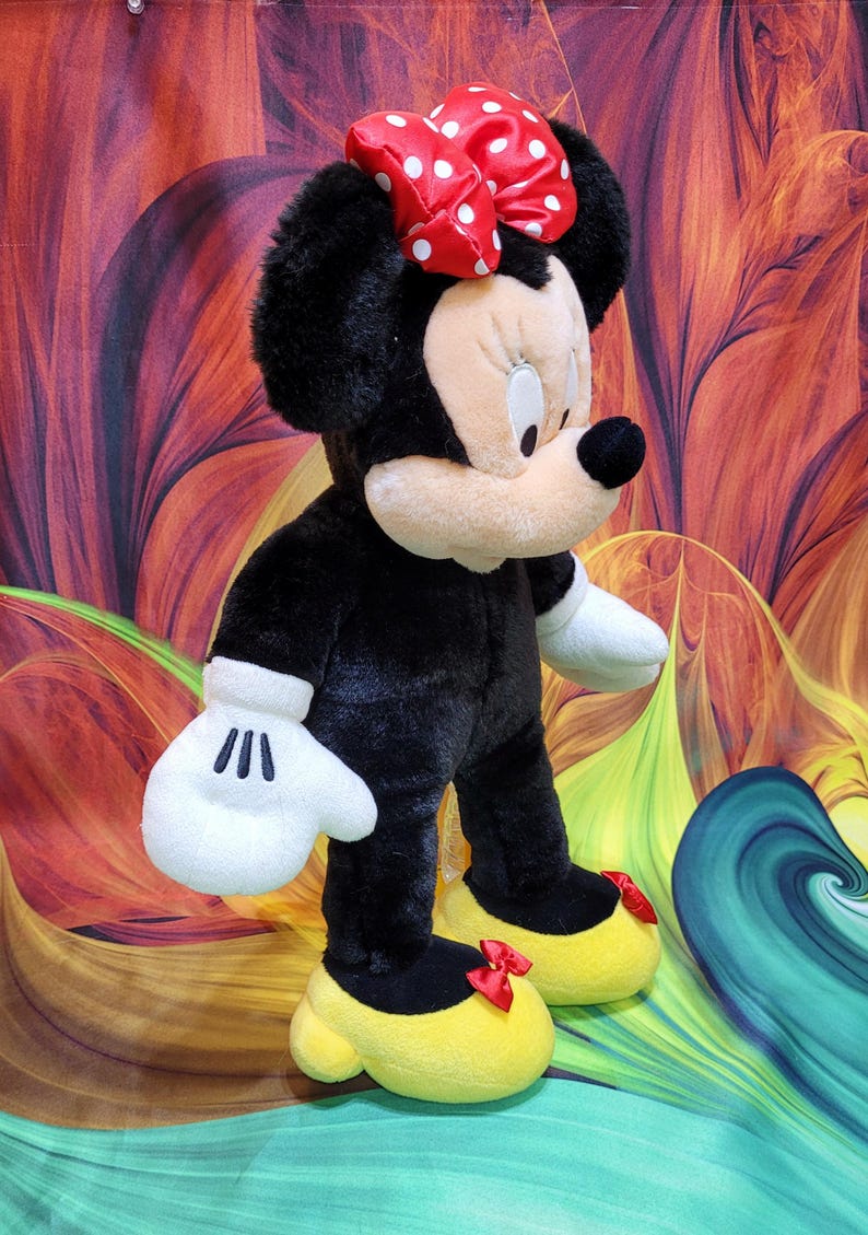 K&ouml;nnte beinhalten: Eine Pl&uuml;schpuppe von Minnie Mouse, mit einem schwarzen K&ouml;rper, wei&szlig;en Handschuhen und gelben Schuhen mit roten Schleifen. Die Puppe hat ein cremefarbenes Gesicht, schwarze Ohren und eine rote Schleife mit Punkten. Die Puppe steht vor einem bunten Hintergrund.