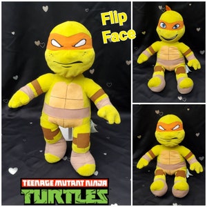 May include: A yellow plush toy of Michelangelo from Teenage Mutant Ninja Turtles. The toy has an orange mask, brown accents, and a flip face feature. The Teenage Mutant Ninja Turtles logo is visible.