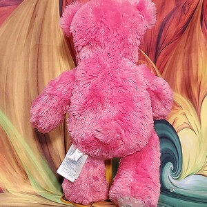 K&ouml;nnte beinhalten: Ein rosa Teddyb&auml;r mit weicher, pelziger Textur. Der B&auml;r wird von hinten betrachtet und zeigt seine Ohren, Arme und Beine. Ein Etikett ist am R&uuml;cken des B&auml;ren befestigt. Der Fu&szlig; des B&auml;ren hat ein kleines Design.